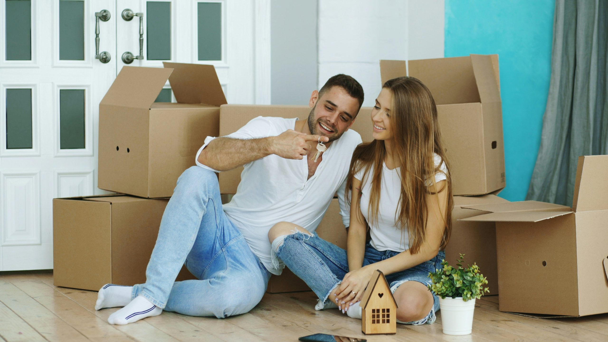 First time home buyers sitting on the floor of their new home surrounded by cardboard moving boxes, holding a small wooden house model next to a potted plant.